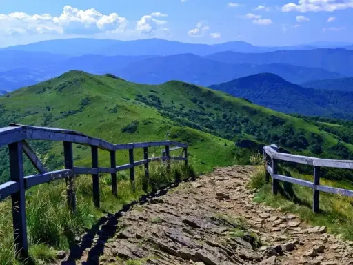Kamienista ścieżka prowadzi przez zielone wzgórza, a drewniane ogrodzenie wyznacza szlak. W oddali widać kolejne, błękitne szczyty w Bieszczadach.