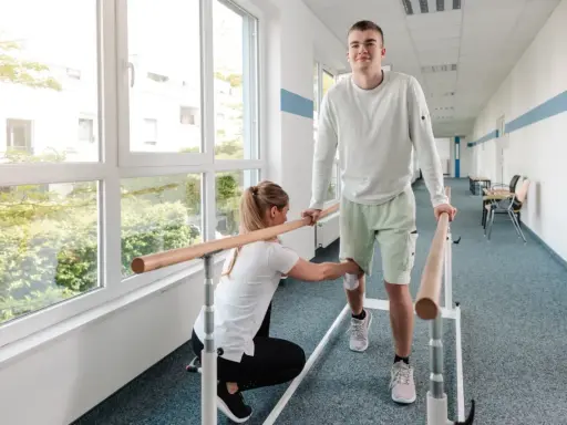 Młody mężczyzna ćwiczy chód przy poręczy, a fizjoterapeutka pomaga mu w rehabilitacji.