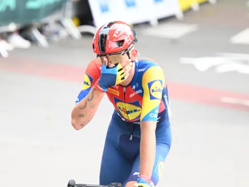 Radfahrer Thibau Nys wischt sich mit der Hand über das Gesicht, trägt einen roten Helm und ein blau-gelbes Trikot.