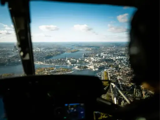 Vy från en helikopter över Stockholm. Staden breder ut sig vid vattnet under en klarblå himmel med moln.