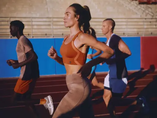 Biegacze na bieżni. Kobieta w pomarańczowym topie i beżowych legginsach, mężczyzna w koszulce i szortach. Buty sportowe z niskim dropem zapewniają komfort.