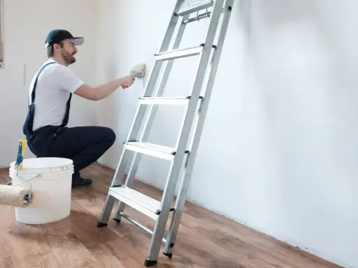Hombre con mono azul y gorra usa brocha para limpiar paredes, junto a escalera y cubo con rodillo.
