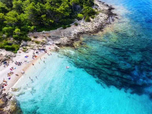 Idealne miejsce, gdzie lecieć w czerwcu: kamienista plaża z turkusową wodą, sosnowy las i opalający się ludzie.