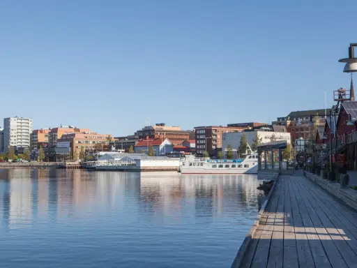 En vacker dag att besöka Luleå. En träspång leder längs vattnet förbi röda byggnader och en vit båt.