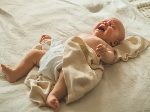 Noworodek leży na plecach, ubrany w pieluszkę, z przykrytymi nóżkami jasnym kocykiem. Jego twarz wykrzywiona jest w płaczu, a z otwartych ust wydobywa się głośne stękanie noworodka podczas snu.