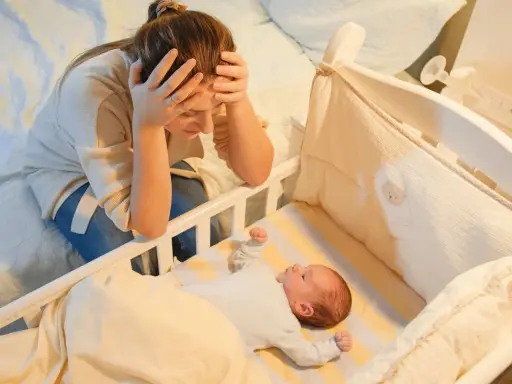 Madre agotada, con las manos en la cabeza, mira a su bebe que se desvela por la noche en su cuna.