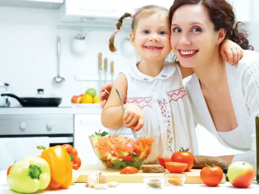 Mama i córka przygotowują sałatkę. W kuchni widać warzywa, oliwę i chleb. To zdrowa żywność, bogata w oligosacharydy, które wspierają florę bakteryjną.
