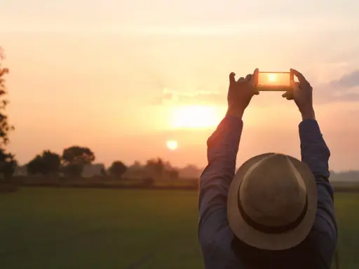 Osoba w kapeluszu fotografuje zachód słońca telefonem z aparatem szerokokątnym.