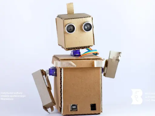 Uroczy robot z kartonu, z oczami-sensorami i kolorowymi kablami, gotowy do nauki i zabawy.