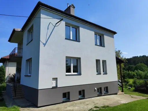 Jasny dom typu kostka z szarym cokołem i balkonem, otoczony zielenią.