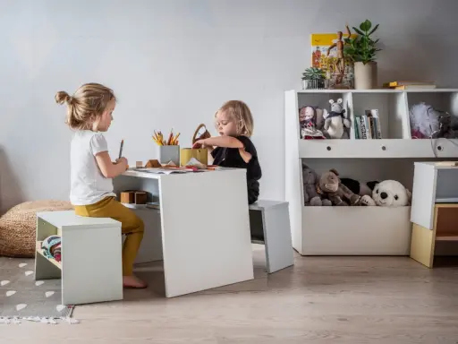 Uroczy kącik dla dziecka w salonie. Dwie dziewczynki rysują przy białym stoliku, otoczone pluszakami i książkami.