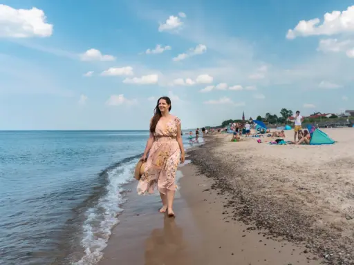 Kobieta w kwiecistej sukience spaceruje brzegiem morza w Sarbinowie. Na plaży widać ludzi odpoczywających na kocach i pod parasolami.