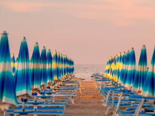 Filetto di ombrelloni a righe blu e bianche sulla spiaggia, con il mare e il cielo rosa all'orizzonte. Un'immagine iconica della riviera adriatica.