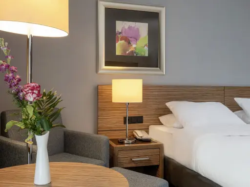 Ein Hotelzimmer mit Bett, Nachttischlampe, Sessel und Blumenstrauß.