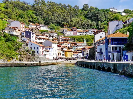 Bunte Häuser schmiegen sich an einen grünen Hang in Cudillero, Spanien. Das türkisfarbene Wasser des Hafens spiegelt die malerische Kulisse wider.