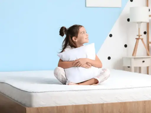 Niña sonriente abraza almohada en un colchón, pensando qué colchon es mejor para niños.