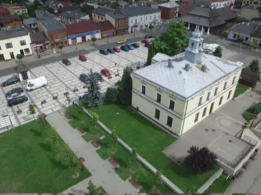 Sławków rynek z lotu ptaka: żółty budynek ratusza, brukowany plac z samochodami i zielony park z ławkami.