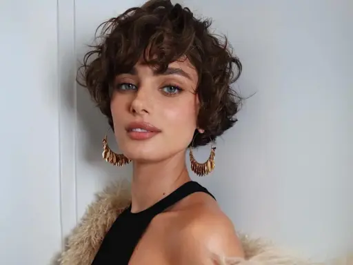 Mujer con un moderno corte shaggy rizado, ojos azules penetrantes y grandes pendientes dorados.