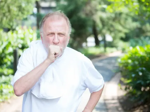 Mann mit COPD hustet im Freien. Sein dicker Bauch ist durch die weiße Kleidung sichtbar.