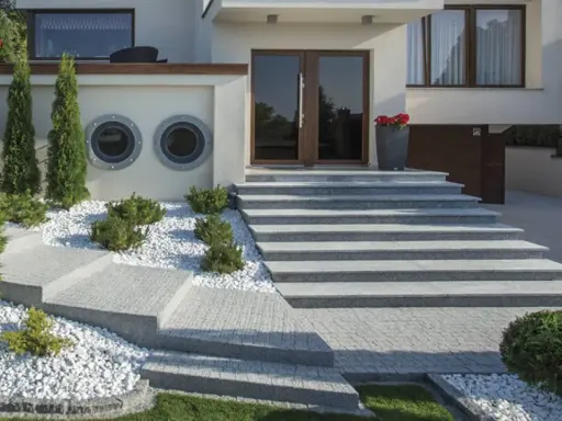 Nowoczesne schody zewnętrzne z beton dekoracyjny, otoczone białym żwirem i zielenią, prowadzące do drzwi wejściowych.