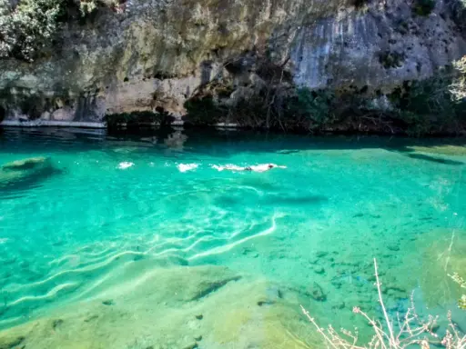 Persona nada en un pou clar de aguas turquesas, bajo un acantilado rocoso.