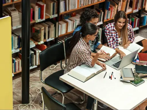 Trzy studentki pracują przy stole w bibliotece wyższej szkoły technicznej, otoczone książkami i laptopem.