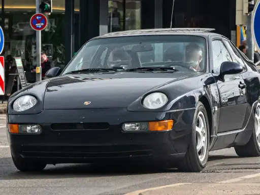 Dunkelblauer 968 Porsche fährt auf der Straße. Ein Mann sitzt am Steuer. Verkehrsschilder sind im Hintergrund zu sehen.