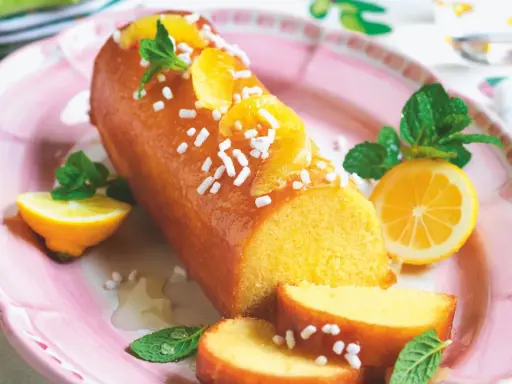 Torta al limone leggerissima, lucida e decorata con fettine d'arancia e zuccherini, servita su un piatto rosa con spicchi di limone e menta.