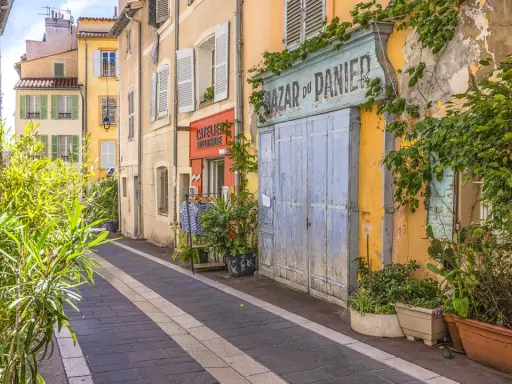 Bunte Häuser und Pflanzen säumen eine Gasse in der Marseille Altstadt. Ein "Bazar du Panier" Schild und blaue Holztüren fallen auf.