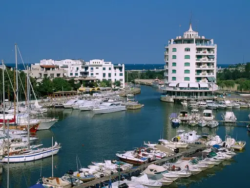 Un vivace porto verde con barche a vela e yacht ormeggiati, edifici bianchi e un edificio circolare a torre.