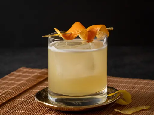 Ein erfrischender Rum Sour mit Eis und Orangenzesten garniert, serviert auf einem Bambusuntersetzer.