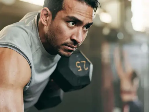 Fokusierter Mann hebt Hantel. Sein Schweiß glänzt. Ein Teil eines 2er Split Trainingsplans.