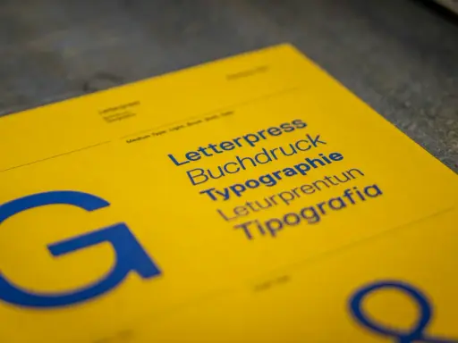 Żółta kartka z niebieskimi napisami: Letterpress, Buchdruck, Typographie, Leturpretun, Tipografia. Duża litera "G" po lewej. To praca typografa.