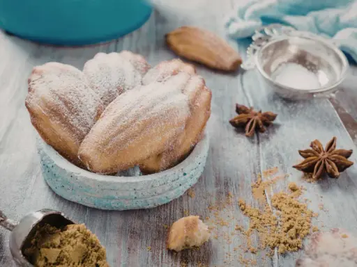 Un bol de biscuits madeleine noisette saupoudrés de sucre glace, avec des étoiles de badiane et une cuillère de poudre sur une table en bois.