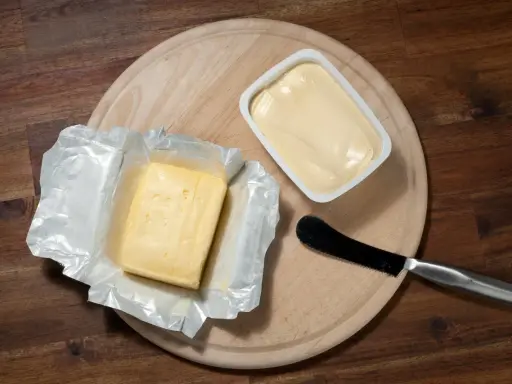 Zwei Butterpackungen, eine in Folie, die andere in einer weißen Schale, liegen auf einem Holzbrett. Eine leichte Butter weniger Fett für den gesunden Genuss.