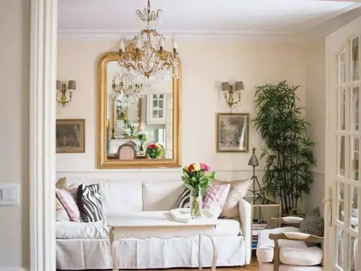 Un salotto con un divano bianco, un grande specchio dorato e un lampadario di cristallo, che evoca un elegante arredamento parigino.