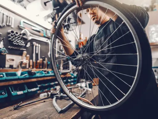 Mechanik sprawdza układy w rowerze, trzymając koło. W tle warsztat z narzędziami.