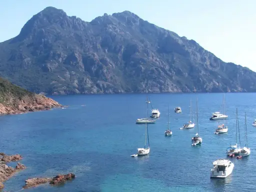Vue d'une baie rocheuse avec plusieurs voiliers et bateaux à moteur en mouillage forain, sous une montagne imposante.