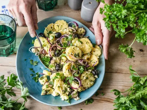 Ein köstlicher mediterraner Kartoffelsalat mit Oliven, roten Zwiebeln und Feta auf einem blauen Teller, garniert mit frischer Petersilie.