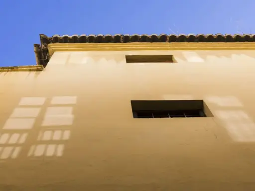 Fachada con enfoscado de cemento color ocre, dos ventanas y la sombra de una reja proyectada sobre el muro.