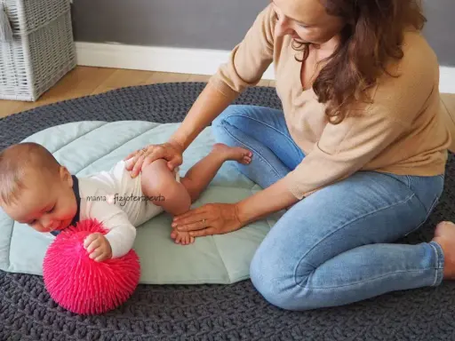 Fizjoterapeutka delikatnie masuje niemowlę, które leży na macie obok różowej piłeczki. Maluch, niczym mała amfibia, ćwiczy pod okiem mamy.