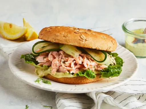 Ein Brötchen mit pulled Lachs, Salat, Gurkenscheiben und einer cremigen Sauce. Dazu Zitronenspalten und ein Dip.