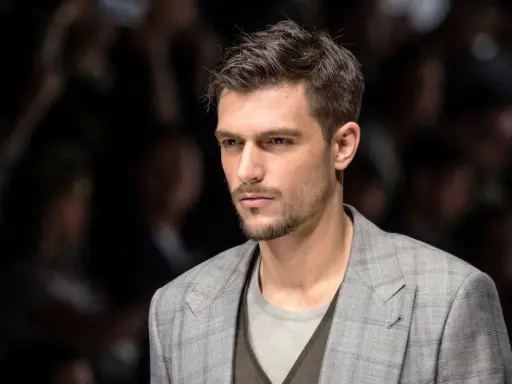 Uomo con capelli spettinati, sguardo intenso, indossa una giacca a quadri grigia.