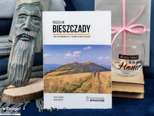 Przewodnik "Ruszaj w Bieszczady" z widokiem na góry i turystów. Obok rzeźba z drewna i świeca z napisem "all you need is in your Home".
