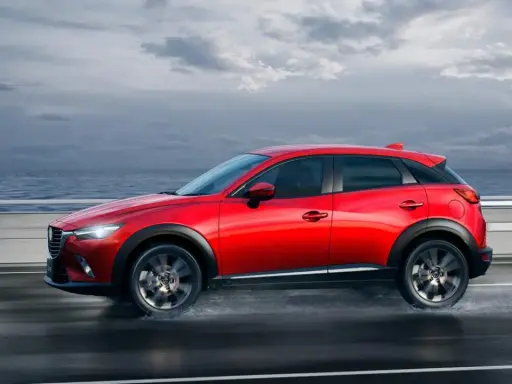 Un Mazda CX-3 rojo circula por una carretera mojada junto al mar. Las medidas del Mazda CX-3 son ideales para la ciudad y la aventura.