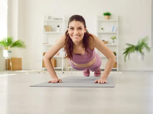 Frau in lila Sportkleidung macht Liegestütze für Yoga Muskelaufbau. Sie lächelt glücklich in die Kamera.