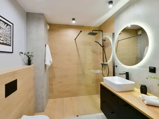 Baño moderno con ducha de suelo continuo, lavabo sobre encimera y espejo circular iluminado.