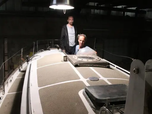 Nicole Van de Kerckhove et un autre homme sur le pont d'un voilier dans un hangar sombre.