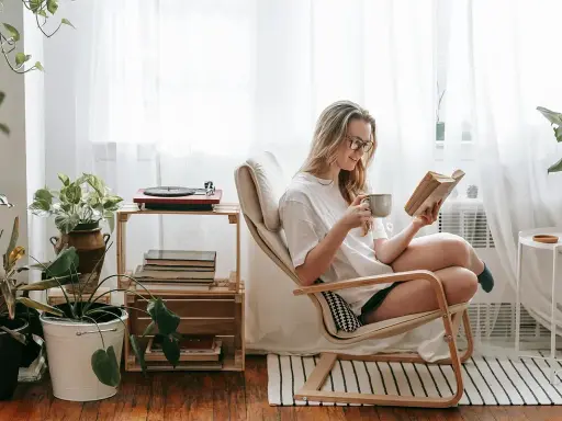 Mujer leyendo en un sillón, taza en mano, rodeada de plantas y libros. Un rincón de lectura con decoración acogedora y un tocadiscos.