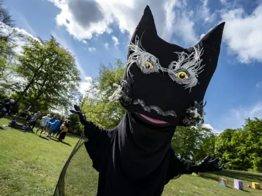 En stor, svart docka som liknar Syrsommarskuggan vinkar glatt i en park med folk i bakgrunden.